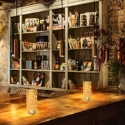 Lámpara cilíndrica recargable de cristal iluminando un elegante espacio exterior, ideal para decoraciones modernas y acogedoras.